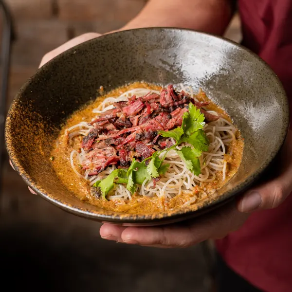 Hands holding out a bowl of rice noodles topped with shredded smoked brisket and coriander