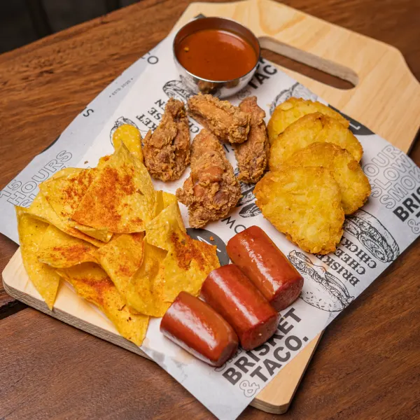 A wooden board of fried chicken, hash browns, seasoned tortilla chips and sausage on LYMA's printed paper
